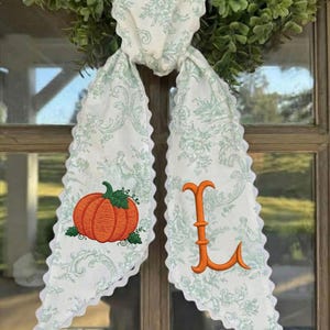 May include: A white fabric door hanger featuring a light green toile pattern with scalloped edges. The left side displays an embroidered orange pumpkin with green leaves, while the right side features a large, embroidered orange letter L.