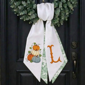 May include: A white fabric door hanger featuring a green eucalyptus wreath at the top. The left panel displays a cluster of three pumpkins in orange, white, and blue-grey. The right panel features a large, orange, decorative letter L.