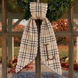 May include: A beige fabric ribbon with a brown, black, and red plaid pattern and white scalloped edges. The ribbon is tied in a knot and features a large, dark brown embroidered letter L on the right tail. The fabric is draped over a window frame.