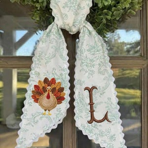 May include: A white fabric scarf with a light green floral toile pattern and scalloped edges. One end features an embroidered turkey with brown, orange, and yellow feathers, while the other end displays a large, brown embroidered letter L.