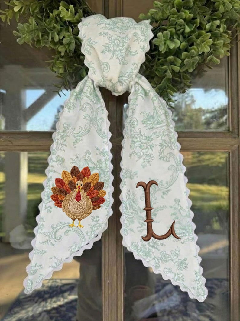 May include: A white fabric scarf with a light green floral toile pattern and scalloped edges. One end features an embroidered turkey with brown, orange, and yellow feathers, while the other end displays a large, brown embroidered letter L.