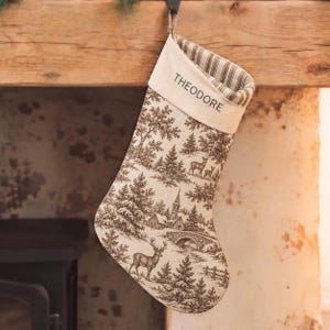 Puede incluir: Un calcetín navideño con un estampado de paisaje invernal en tonos marrón y crema estilo toile, con ciervos, árboles y un puente de piedra. El puño es de tela color crema con el nombre THEODORE impreso en negro y forro interior a rayas.