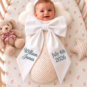 May include: A baby wrapped in a cream-coloured swaddle blanket featuring a large white bow. The left tail of the bow is embroidered with the text William James, and the right tail features the text July 4th 2026 in dark teal lettering.
