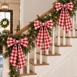 May include: A staircase railing decorated with a lush green pine garland featuring small warm white string lights and three large red and white buffalo check fabric bows attached at intervals along the wooden banister.