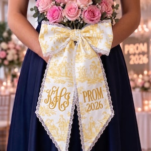 May include: A bouquet of pink roses wrapped in a white and yellow toile fabric ribbon with lace trim. The ribbon features a yellow toile pattern, a gold embroidered monogram with the letters KHS, and the gold embroidered text PROM 2026.