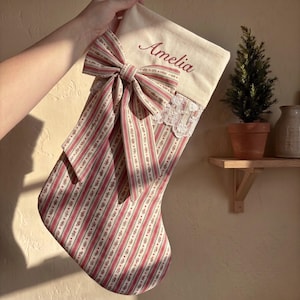 以下が含まれることがあります： 赤色の筆記体で「Amelia」と刺繍されたクリーム色のカフが付いたクリスマスストッキング。ストッキング本体は赤と緑の縦縞と花柄の生地で作られており、大きな共布のリボンとレースの装飾が施されています。