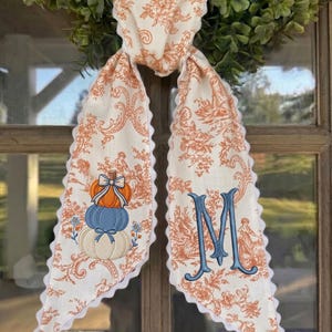 May include: A long, cream-colored fabric scarf featuring an orange toile floral pattern and white scalloped edges. One side displays a stack of three pumpkins in orange, blue, and cream, while the other side features a large, embroidered blue letter M.