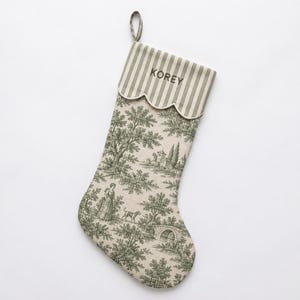 Puede incluir: Un calcetín navideño de tela beige con un patrón toile de jouy verde que representa un paisaje con una persona, un perro y un puente. El puño es de rayas verdes y blancas con un borde festoneado y el nombre KOREY bordado en verde.