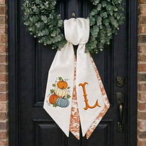 May include: A green eucalyptus wreath featuring a hanging white fabric sash. The left side of the sash displays a stack of three pumpkins in orange, white, and blue. The right side of the sash features a large, orange, decorative letter L.