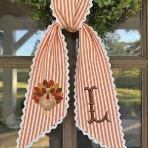 May include: A decorative fabric door hanger featuring orange and white vertical stripes with white scalloped lace trim. One side displays an embroidered turkey with colorful feathers, while the other side features a large, brown embroidered letter L.