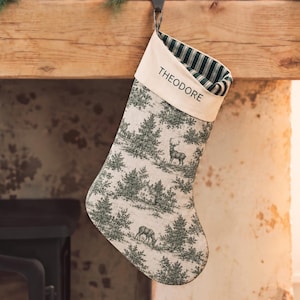 Op de afbeelding: Een kerstkous met een crèmekleurige boord waarop de naam THEODORE in het zwart is gedrukt. Het lichaam van de kous toont een donkergroen en crème toile-patroon van herten, bomen en een huis, met een donkergroene gestreepte voering aan de binnenkant.