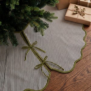 May include: A circular tree skirt featuring a black and white vertical pinstripe pattern with a scalloped edge trimmed in olive green fabric. The skirt includes two olive green fabric ties for closure, positioned beneath a green pine tree branch.