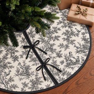 May include: A circular Christmas tree skirt featuring a cream-coloured fabric with a black toile print depicting pastoral scenes of people and animals among trees. The skirt has a black fabric border and two black ribbon ties for closure.