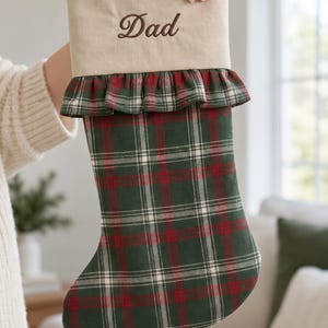 May include: A Christmas stocking featuring a beige cuff embroidered with the word Dad in brown thread. Below the cuff is a ruffled trim in a green, red, and white plaid pattern, which continues down the body of the stocking in the same plaid.