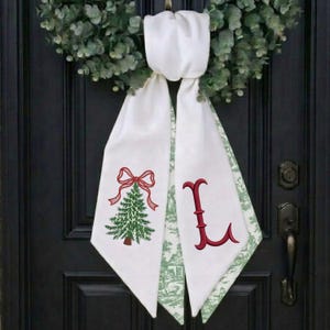 May include: A white fabric door hanger featuring a green embroidered Christmas tree topped with a red bow on one side and a large red embroidered letter L on the other, with a green and white toile pattern on the reverse side of the fabric.