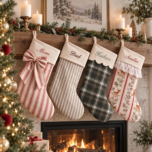 Puede incluir: Cuatro calcetines navideños colgantes con puños de color crema bordados con los nombres Mom, Dad, Hudson y Amelia. Los calcetines presentan rayas rojas y blancas con un lazo, rayas grises, un patrón de cuadros verde oscuro y un estampado floral de ciervos.