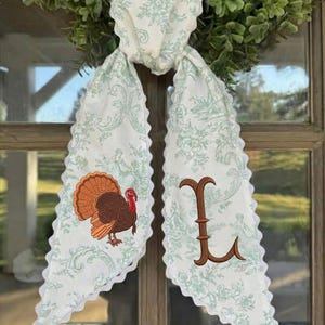 May include: A white fabric scarf with a light green floral toile pattern and scalloped edges. One side features an embroidered brown turkey with a red wattle, and the other side displays a large, brown embroidered letter L in a serif font.