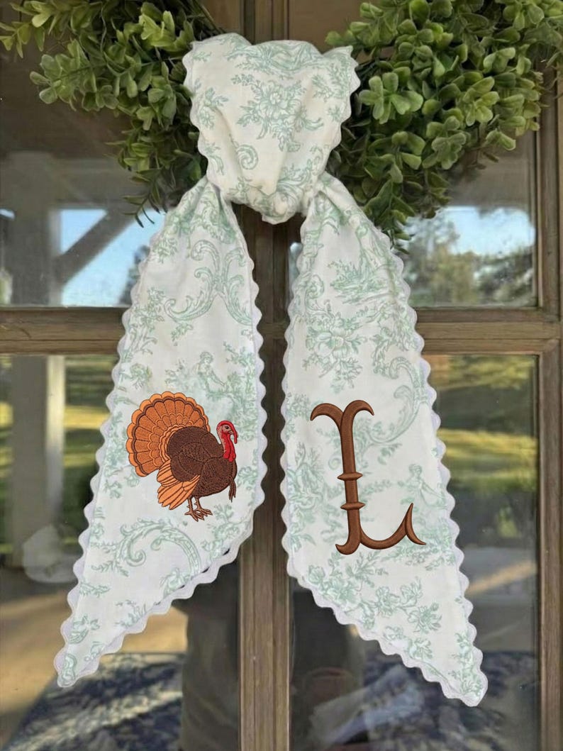 May include: A white fabric scarf with a light green floral toile pattern and scalloped edges. One side features an embroidered brown turkey with a red wattle, and the other side displays a large, brown embroidered letter L in a serif font.