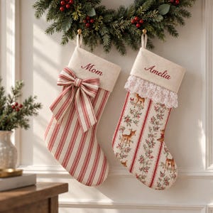 Puede incluir: Dos calcetines navideños con puños color crema bordados con los nombres Mom y Amelia. Uno tiene rayas verticales rojas y blancas con un lazo a juego, mientras que el otro presenta un borde de encaje blanco y un estampado de ciervos y acebo.
