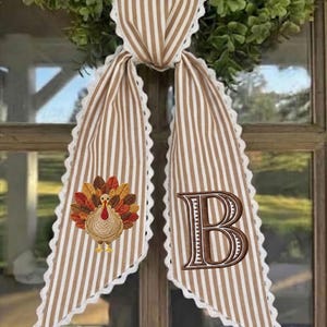 May include: A decorative fabric door hanger featuring a tan and white vertical striped pattern with white scalloped lace trim. One side displays an embroidered turkey with colorful feathers, while the other side features a large embroidered letter B.