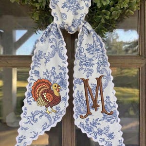May include: A white fabric scarf with a blue toile floral pattern and scalloped edges. One side features an embroidered brown turkey with orange and red feathers, while the other side displays a large, embroidered brown letter M monogram.
