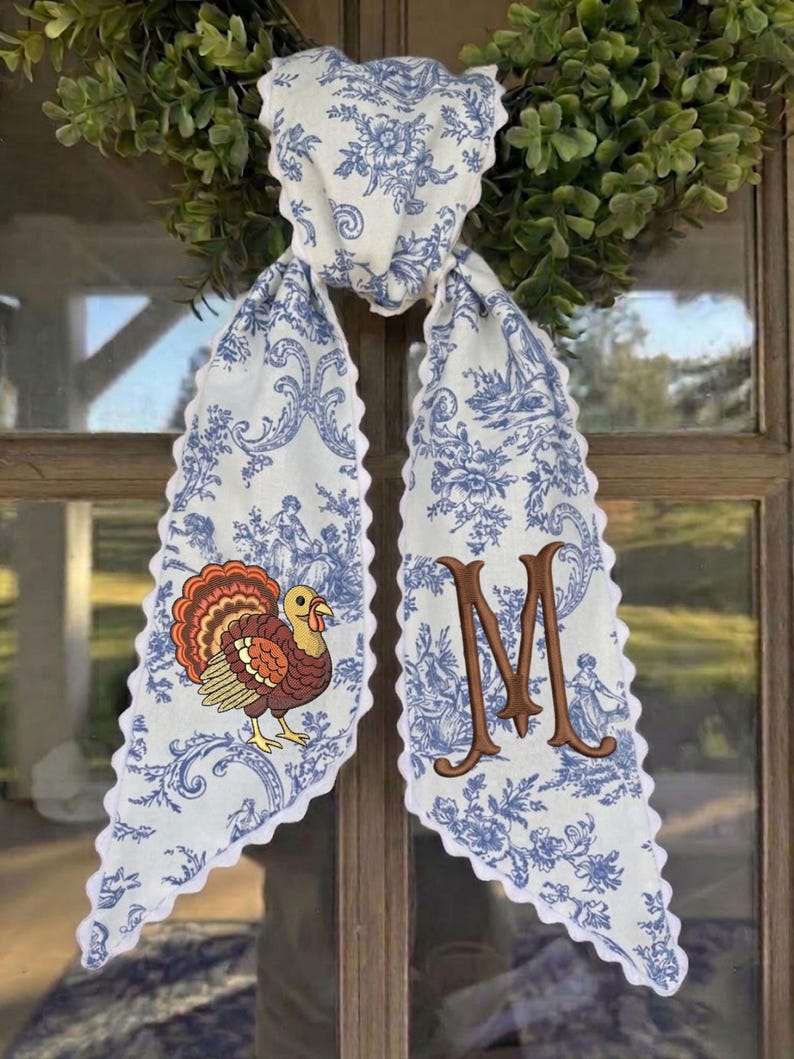 May include: A white fabric scarf with a blue toile floral pattern and scalloped edges. One side features an embroidered brown turkey with orange and red feathers, while the other side displays a large, embroidered brown letter M monogram.