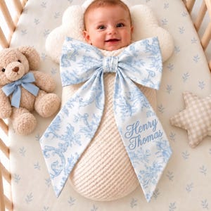 Könnte beinhalten: Ein Baby in einem cremefarbenen Wickeltuch mit einer großen blau-weißen Schleife im Toile-de-Jouy-Muster, auf der der Name Henry Thomas gestickt ist, dazu ein hellbrauner Teddybär mit blauer Schleife und ein beiges, sternförmiges Kissen.