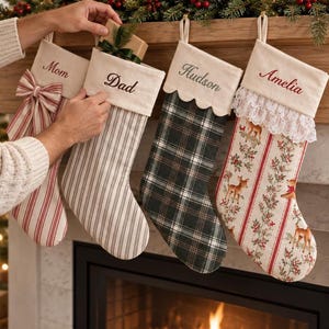 Puede incluir: Cuatro calcetines navideños personalizados colgados en fila. El primero tiene rayas rojas y un lazo, el segundo rayas grises y el nombre Dad, el tercero un patrón de cuadros verdes y el nombre Hudson, y el cuarto un estampado de ciervos y el nombre Amelia.