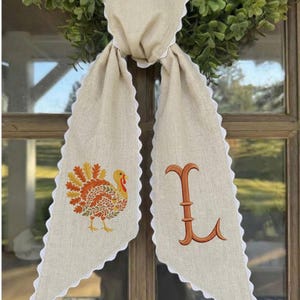 May include: A beige linen-like fabric door hanger featuring a scalloped white trim edge. The left side displays an embroidered turkey with orange and yellow leaf-patterned feathers, while the right side features a large, embroidered orange letter L.