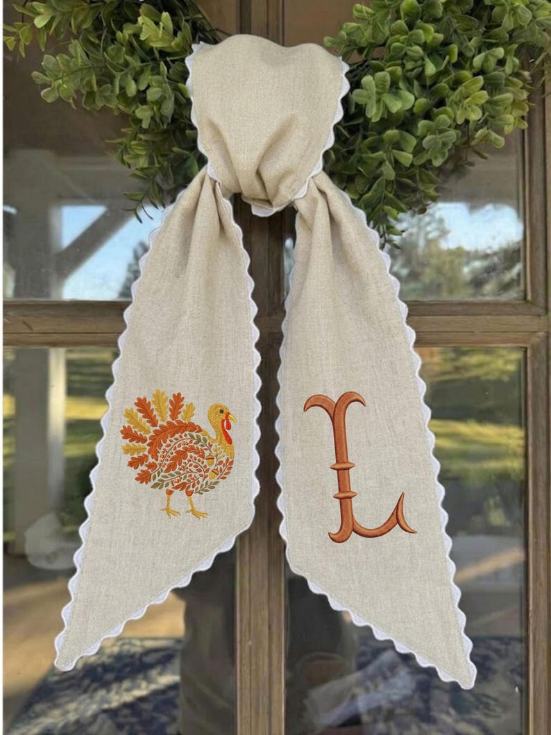 May include: A beige linen-like fabric door hanger featuring a scalloped white trim edge. The left side displays an embroidered turkey with orange and yellow leaf-patterned feathers, while the right side features a large, embroidered orange letter L.