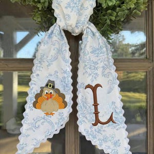 May include: A white fabric scarf with a light blue floral toile pattern and scalloped edges. One side features an embroidered turkey wearing a pilgrim hat, while the other side displays a large, brown, decorative embroidered letter L.