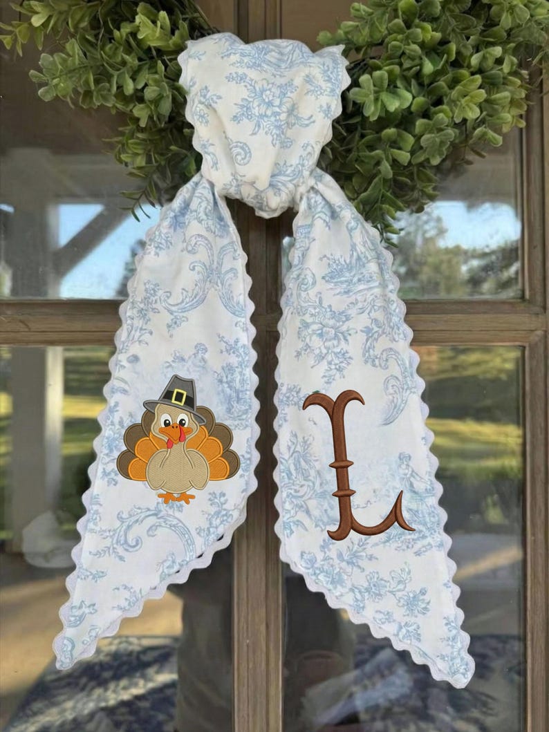May include: A white fabric scarf with a light blue floral toile pattern and scalloped edges. One side features an embroidered turkey wearing a pilgrim hat, while the other side displays a large, brown, decorative embroidered letter L.