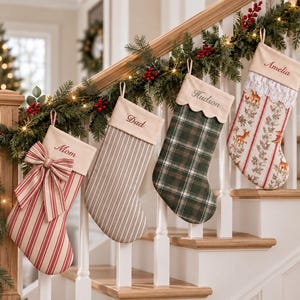 Puede incluir: Cuatro calcetines de Navidad colgados en fila. El primero es de rayas rojas y blancas con un lazo grande y bordado 'Mom'. El segundo es beige con rayas marrones finas y 'Dad'. El tercero es de cuadros verdes con 'Hudson'. El cuarto tiene estampado de ciervos, encaje y 'Amelia'.