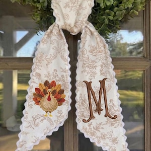 May include: A cream-coloured fabric door hanger featuring a tan floral toile pattern and a scalloped white edge. The left side displays an embroidered turkey with colourful feathers, while the right side features a large, brown embroidered letter M.