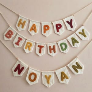 May include: A festive birthday banner with the words "HAPPY BIRTHDAY NOYAN" spelled out in colorful, embroidered letters. The letters are in shades of orange, red, yellow, and green, sewn onto white pennants, strung together with a neutral cord.