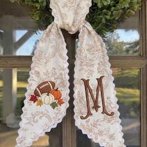 May include: A cream-coloured fabric door hanger featuring a tan toile floral pattern and scalloped edges. One side displays an embroidered football, a white pumpkin, an orange pumpkin, and autumn leaves. The other side features a large brown letter M.