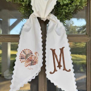 May include: A white fabric scarf with scalloped edges featuring an embroidered turkey on one side and a large brown letter M on the other. The scarf is tied in a knot and hangs down, displaying detailed autumn-colored leaf patterns on the turkey.