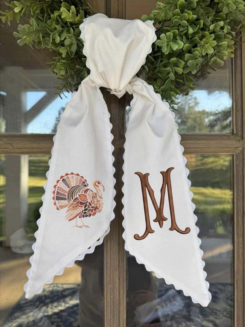 May include: A white fabric scarf with scalloped edges featuring an embroidered turkey on one side and a large brown letter M on the other. The scarf is tied in a knot and hangs down, displaying detailed autumn-colored leaf patterns on the turkey.