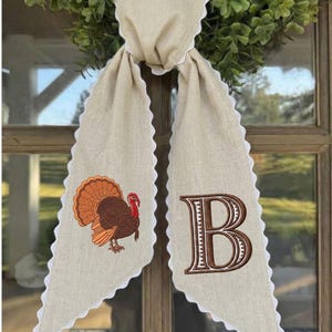May include: A beige linen-like fabric door hanger featuring a scalloped white edge. One side displays an embroidered brown and orange turkey, while the other side features a large, embroidered brown letter B with white detailing.