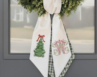 May include: White Christmas door sash with a green Christmas tree and red bow embroidery on one side and a monogram on the other. The sash is draped over a green wreath, creating a festive holiday decoration.