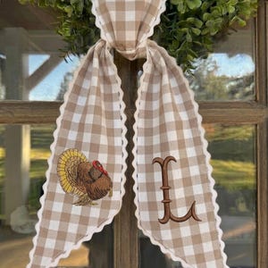 May include: A beige and white gingham fabric ribbon with scalloped edges, tied in a knot. The left tail features an embroidered turkey with brown, yellow, and red feathers, while the right tail displays a large, brown embroidered letter L.