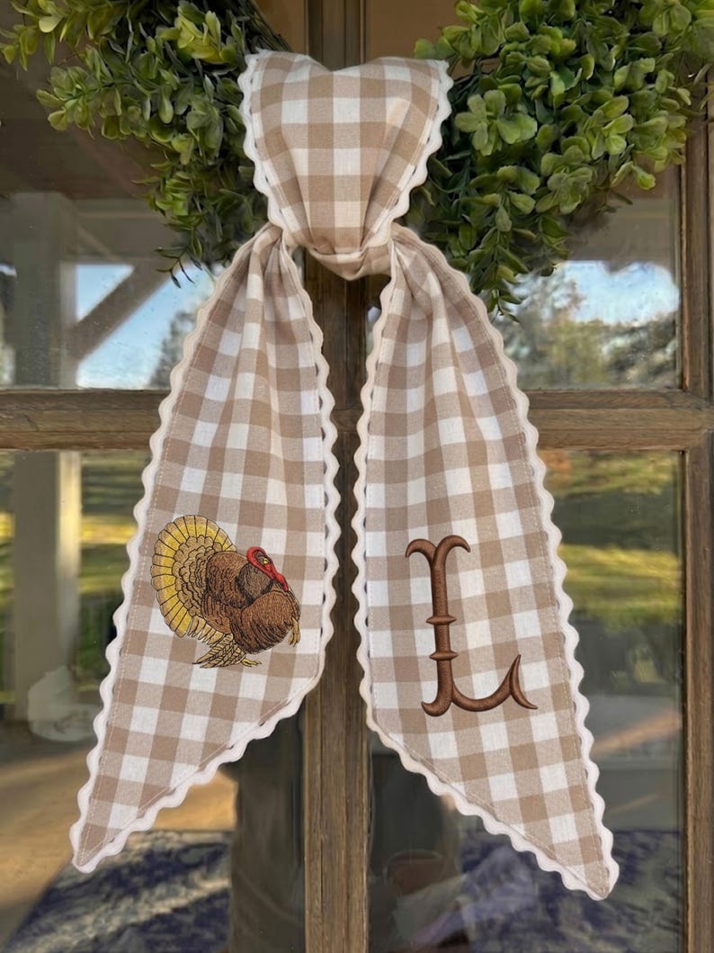 May include: A beige and white gingham fabric ribbon with scalloped edges, tied in a knot. The left tail features an embroidered turkey with brown, yellow, and red feathers, while the right tail displays a large, brown embroidered letter L.