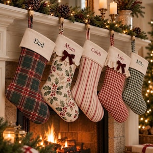 May include: Five Christmas stockings hanging in a row, each with a cream-coloured cuff personalised with the names Dad, Mom, Caleb, Lily, and Milo. The stockings feature various patterns including green and red plaid, holly leaves, red stripes, and red or green gingham checks.