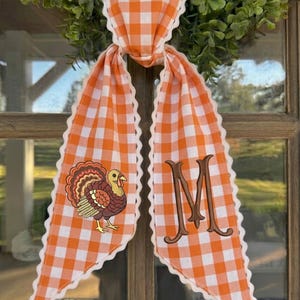 May include: A decorative fabric door hanger featuring an orange and white gingham pattern with scalloped white edges. One side displays an embroidered brown turkey, while the other side features a large, embroidered brown letter M monogram.
