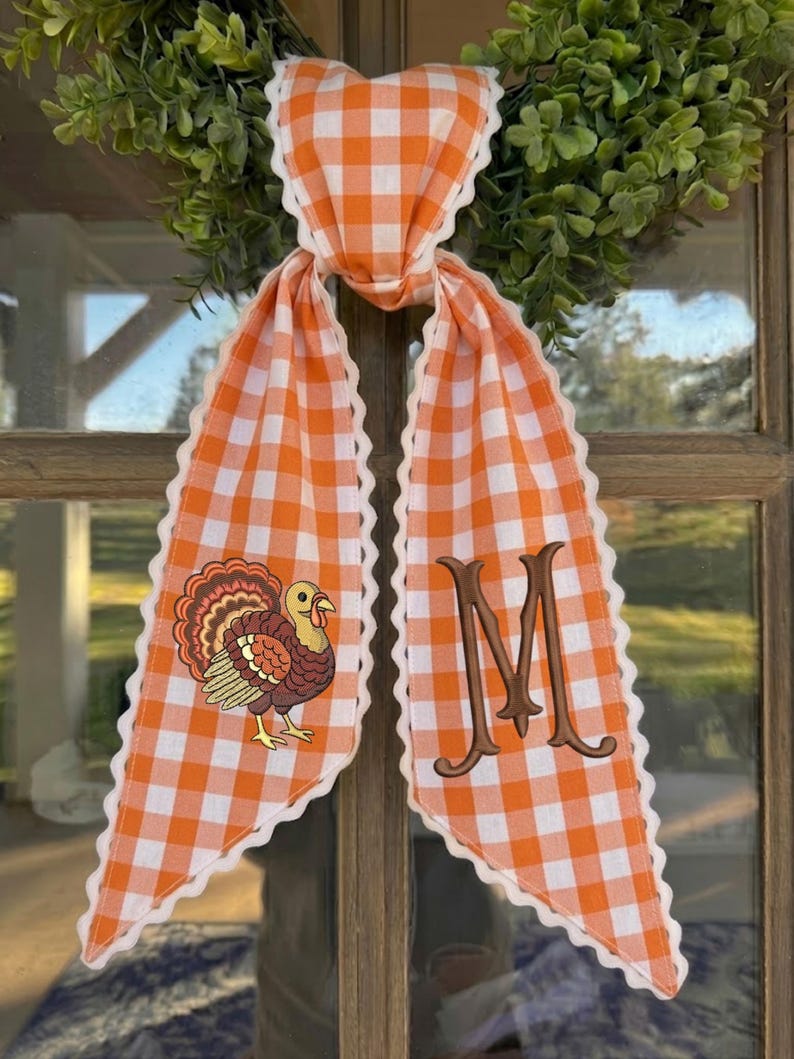 May include: A decorative fabric door hanger featuring an orange and white gingham pattern with scalloped white edges. One side displays an embroidered brown turkey, while the other side features a large, embroidered brown letter M monogram.