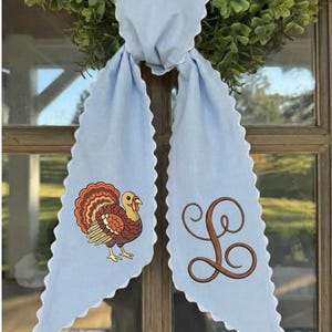 May include: A light blue fabric door hanger with scalloped white edges. The left side features an embroidered turkey with brown, orange, and red feathers. The right side features a large, brown, cursive embroidered letter L monogram.