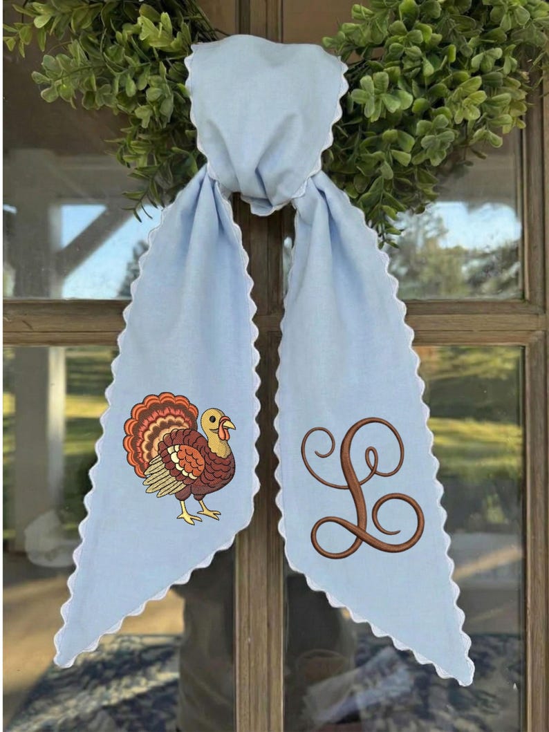 May include: A light blue fabric door hanger with scalloped white edges. The left side features an embroidered turkey with brown, orange, and red feathers. The right side features a large, brown, cursive embroidered letter L monogram.