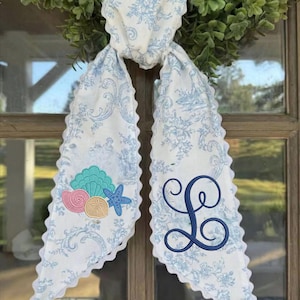 May include: A white fabric hair accessory with a light blue floral toile pattern and scalloped edges. One side features an embroidered design of a teal seashell, a pink seashell, a tan sand dollar, and a blue starfish. The other side has a navy blue monogram letter L.