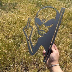 May include: A black metal alien silhouette with a raised middle finger. The alien has large oval eyes and a small mouth. The metal cutout is held against a background of tall, dry grass.