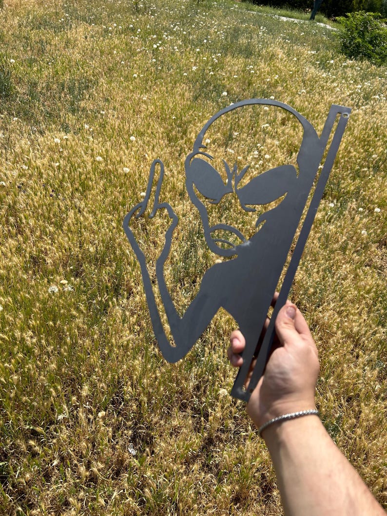 May include: A black metal alien silhouette with a raised middle finger. The alien has large oval eyes and a small mouth. The metal cutout is held against a background of tall, dry grass.